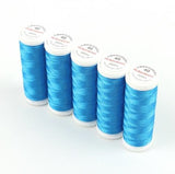 Fünf Spulen ACKERMANN®-Stickgarn Stärke 40 (Farbe 2854 blau, 5 × 260 m) sind diagonal auf einer weißen Fläche angeordnet. Jede Spule ist mit einer weißen Kappe versehen, auf der roter und blauer Text zu sehen ist.