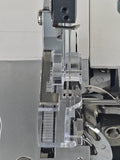 Nahaufnahme des Nadelbereichs einer Nähmaschine mit zwei Nadeln und einem Nähfuß – dem „Klarsichtfuß für Gritzner 788 Overlock (Original)“ von ACKERMANN® – als Teil des Original-Zubehörs, zusammen mit sichtbaren Metallkomponenten, durchsichtigen Kunststoffführungen und mechanischen Teilen.